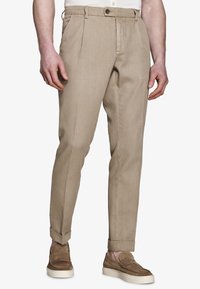Hombre vestido con pantalones beige plisados con puños, combinados con mocasines de ante marrón y una camisa blanca, de pie frente a un fondo liso.