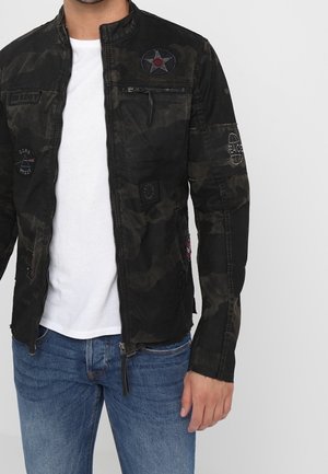 Mężczyzna w czarnej kurtce moro z naszywkami, białej koszulce i niebieskich dżinsach, stojący na jednolitym tle.