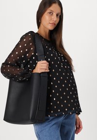 Borsa tote nera in pelle con forma strutturata, ampia tracolla regolabile e texture liscia, tenuta da una modella in una blusa a pois.