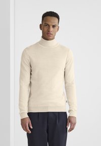 Jersey de cuello alto beige hecho de un suave tejido de punto, con mangas largas, cuello acanalado y un ajuste entallado. Combinado con pantalones oscuros.