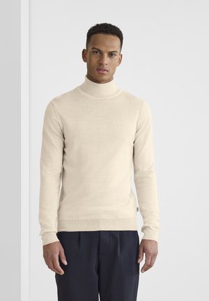 Beige turtleneck sweater made of soft knit fabric, featuring long sleeves, ribbed collar, and a slim fit. Paired with dark pants.