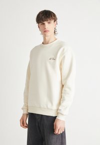 Sudadera color crema de tejido suave, con puños y cuello acanalados, y un pequeño logo bordado en el pecho.