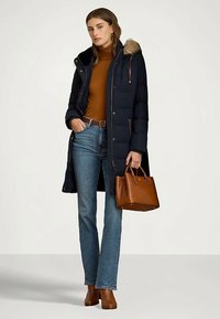 Γυναίκα με ναυτικό φουσκωτό παλτό με γούνινη κουκούλα, καφέ τρουακάρ, μπλε τζιν, καφέ μποτάκια μέχρι τον αστράγαλο και κρατάει μια καφέ δερμάτινη τσάντα.