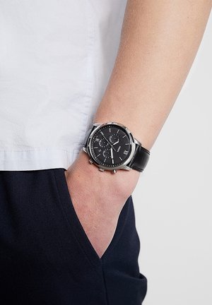 Csuklón fekete bőrszíjas óra fekete számlappal és ezüst tokkal, kéz laza módon sötét nadrág zsebében, fehér ing háttérrel.