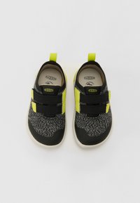 Sneaker nere e grigie con texture, dettagli in neon giallo, cinturini in velcro, suole in gomma e tomaia in tessuto, progettate per comfort e flessibilità.