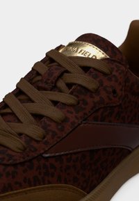 Κοντινό πλάνο καφέ αθλητικού παπουτσιού με λεοπάρ σχέδιο, καφέ κορδόνια, χρυσή ετικέτα στη γλώσσα και λεπτομέρεια με σκούρα καφέ ρίγα.