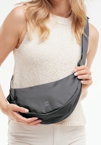 Sac bandoulière en tissu gris avec une forme incurvée et une sangle ajustable, affichant un logo blanc. Texture lisse avec un design moderne.