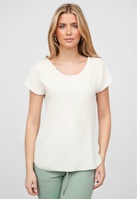 Kortermet lys beige topp med dyp utringning, myk tekstur og litt buet nederkant, kombinert med grønne bukser.