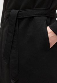 Μαύρη ρόμπα από ύφασμα με ζώνη δεμένη στη μέση, που διαθέτει πλαϊνή τσέπη. Ομαλή υφή και απλός σχεδιασμός, που τονίζει τη λειτουργικότητα και την άνεση.