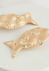 Dos objetos decorativos en forma de pez dorado con una superficie texturizada, colocados sobre una base redonda blanca. Finamente elaborados, con patrones irregulares.