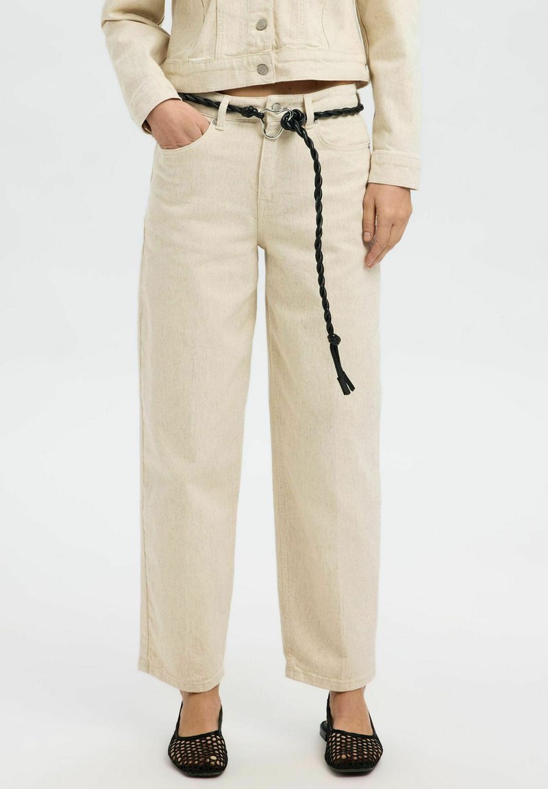 Beige wijde broek gemaakt van denim met een gevlochten zwarte riem, met zakken en een subtiele textuur. Gecombineerd met zwarte mesh schoenen.