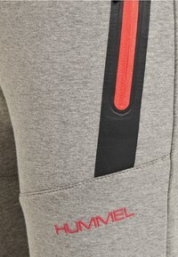 Grå sweatpants med en texturerad tyg, svart sidopanel med en röd dragkedja och en röd logotyp på framsidan.