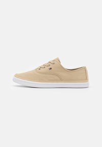Zapatilla baja de lona beige con suela de goma blanca, parte delantera con cordones y pequeño logo rojo, blanco y azul en el lateral.