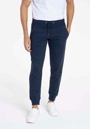 Pantalons de jogging bleu marine en tissu doux, avec une taille élastique munie de cordons de serrage, des poignets côtelés et des poches latérales. Portés avec des baskets blanches.