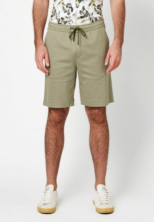 Knielange grüne Shorts aus weichem Stoff mit elastischem Bund, verstellbarem Kordelzug und Seitentaschen. Kombiniert mit weißen Sneakers.