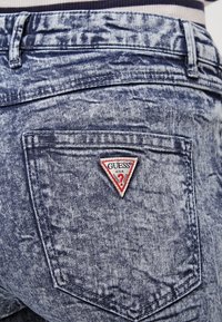 Dżinsy typu denim w jasnoniebieskim, wyblak�łym kolorze. Posiadają trójkątną, czerwoną naszywkę z logo "GUESS" na tylnej kieszeni. Wzdłuż kieszeni widoczne przeszycia.