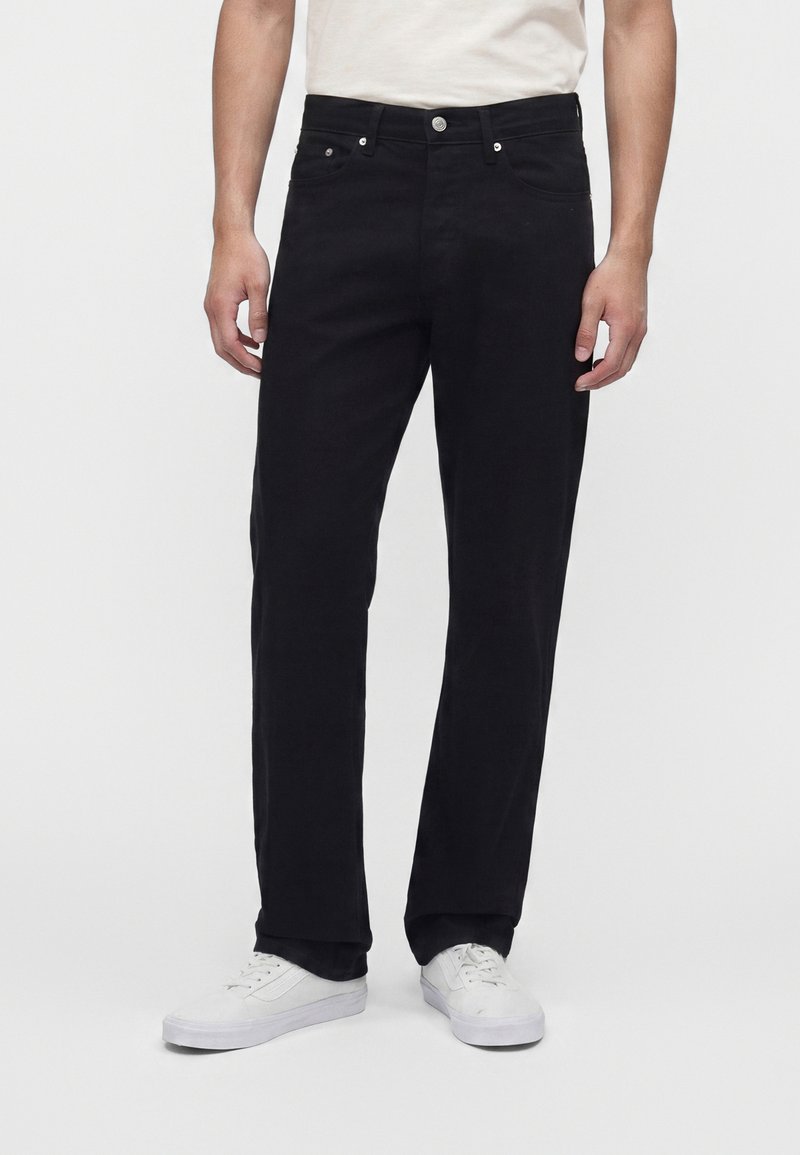 Uomo che indossa pantaloni neri dritti e sneaker bianche in piedi davanti a uno sfondo bianco uniforme.