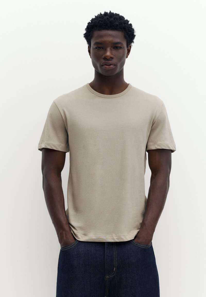 Jeune homme aux cheveux courts et bouclés portant un t-shirt beige à manches courtes et un jean bleu foncé, debout les mains dans les poches.