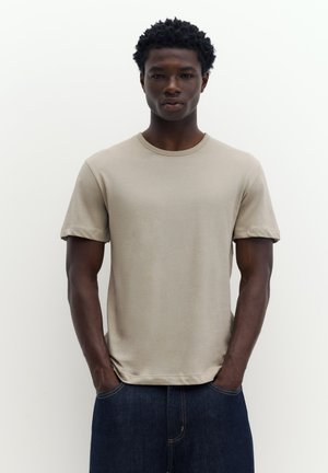 Jeune homme aux cheveux courts et bouclés portant un t-shirt beige à manches courtes et un jean bleu foncé, debout les mains dans les poches.