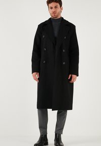 Melnas vilnas apmetnis ar dubulto pogu dizainu, lieliem atloka galiem un melnām pogām. Uzvilkts virs pelēka turtlenecka un pelēkām biksēm.