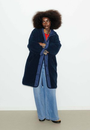 Manteau teddy bleu marine avec des boutons sur le côté, superposé à une veste intérieure matelassée, associé à un jean en denim à jambes larges et des chaussures noires.