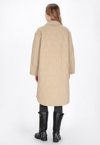 Vatteret beige lang frakke med en bred krave og mønstret tekstur, parret med sorte knæhøje støvler med spænde accenter.