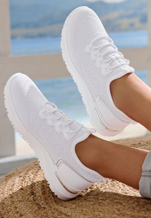 Weiße Netzsneaker mit einer strukturierten Oberseite, flachen Schnürsenkeln und einem silbernen Akzent an der Ferse. Sie haben eine dicke weiße Sohle und werden auf einem gewebten Teppich präsentiert.