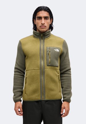 Giovane uomo con giacca in pile verde oliva e verde scuro con zip frontale completa e tasche con zip, in piedi davanti a uno sfondo chiaro e uniforme.