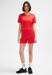 Rött atletiskt set med kortärmad topp och shorts i fukttransporterande tyg. Logodetalj på bröstet. Bärs med svarta skor och vita strumpor.