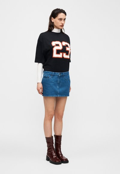 Γυναίκα που φοράει μαύρο φαρδύ μπλουζάκι με τον αριθμό 23, λευκό μακρυμάνικο πουκάμισο από μέσα, μπλε τζιν μίνι φούστα και καφέ μποτάκια μέχρι τον αστράγαλο.