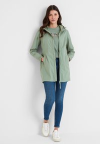 Light green, water-resistant jacket with a hood, elastic cuffs, two pockets, and a front zipper. Paired with a grey t-shirt and blue jeans.