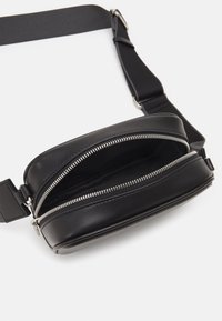 Černá kožená krosbody taška s zipovými přihrádkami, plochým tvarem, nastavitelným textilním popruhem a stříbrnými kovovými detaily.