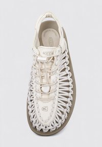 Zapatilla deportiva de color beige claro con parte superior de tejido trenzado, presenta una suela gruesa gris y un sistema de cordones elásticos para un ajuste seguro. Lados texturizados y marca.