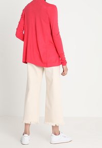 Helles pinkes Langarmoberteil, lockere Passform; kombiniert mit off-white weiten Hosen, mit fransigem Saum. Weiße Sneaker runden das Outfit ab.