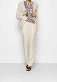 Beige stickad cardigan över en vit leopardmönstrad blus, parat med ljusa, flödande krämfärgade byxor och svarta halksandaler.