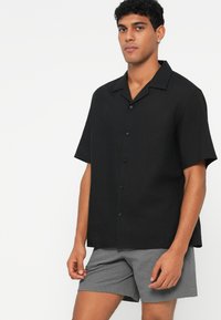 Black short-sleeve button-up shirt with a relaxed fit, featuring a notched collar and subtle texture. Worn with gray textured shorts.