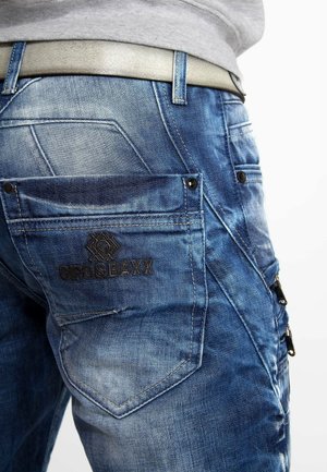 Jean en denim bleu avec une finition délavée, doté d'une poche plaquée avec un logo en relief, de coutures contrastées et d'un détail de poche zippée en métal.