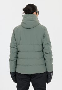 Giacca a vento in verde bosco con cappuccio, caratterizzata da un design trapuntato e polsini elastici. Guanti isolanti neri visibili ai polsi.