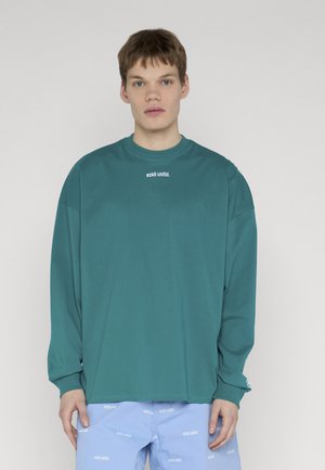 Junger Mann trägt ein tealfarbenes Langarmshirt und hellblaue Shorts mit weißen "eckō unltd." Logos auf schlichtem hellgrauem Hintergrund.