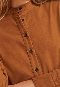 Camicia in velluto a coste marrone con scollatura rotonda, bottoni neri e una sottile trama a coste, dal fit rilassato e maniche lunghe.