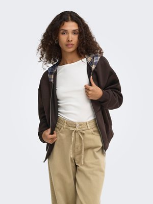 Donna con capelli ricci che indossa una felpa con cappuccio marrone scuro con zip e fodera a quadri, camicia bianca e pantaloni beige con coulisse, che guarda in avanti.