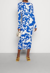 Robe à manches longues blanche avec un motif abstrait de vagues bleues, dotée de fronces sur le côté et d'une longueur midi. Associée à des sandales jaunes.
