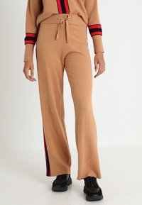 Beżowe dzianinowe spodnie z rozszerzanymi nogawkami, wyposażone w ściągacz ze sznurkiem w pasie oraz czerwone paski po bokach. Miękka faktura, casualowy design.