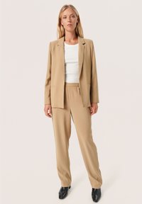 Beige přizpůsobený blazer a odpovídající kalhoty, vyrobené z hladké tkaniny. Blazer má zapínání na jeden knoflík a dvě přední kapsy. Lze kombinovat s upnutou bílou košilí.
