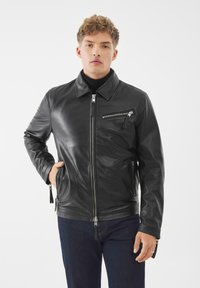 Černá kožená bunda s zipovým zapínáním, špičatým límcem a bočními kapsami. Obsahuje malou kapsu na zip na hrudi a má hladkou texturu. Pod ní jsou nošeny modré džíny.