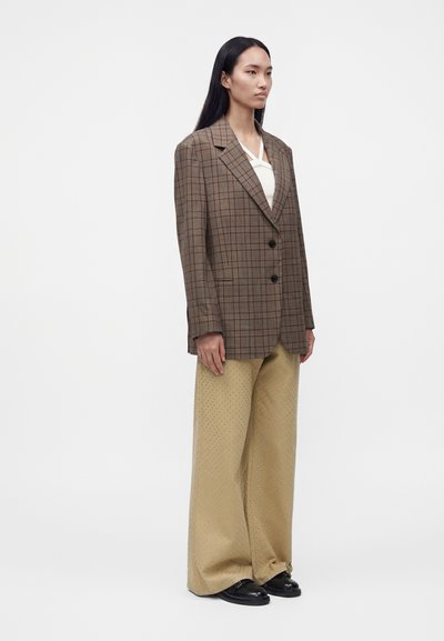 Femme aux longs cheveux noirs portant un blazer marron à carreaux, un haut blanc, un pantalon beige large avec de petits pois et des chaussures noires, debout de côté.