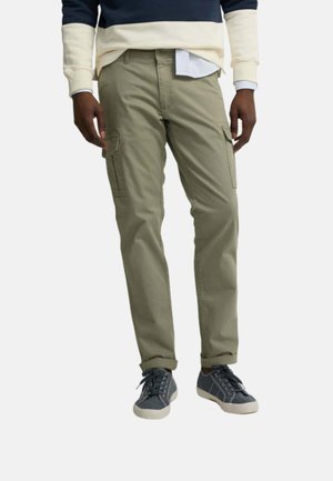 Hombre con pantalones cargo oliva remangados, jersey azul marino y crema, camisa blanca y zapatillas deportivas grises, de pie frente a un fondo liso.