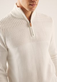 Suéter blanco con cremallera, que presenta un cuello acanalado, un patrón texturizado en la parte superior y un cuerpo liso en la parte inferior. Cremallera con un acento de cuero en el cuello.