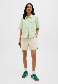 Camisa corta de manga verde claro con cierre de botones y un bolsillo en el pecho, combinada con pantalones cortos beige y zapatillas verdes con detalles en blanco.