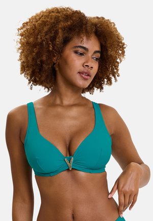 Femme aux cheveux bouclés portant un haut de bikini bleu sarcelle avec une boucle en forme de V dorée, ajustant le côté avec sa main droite, regardant vers le bas et à droite.